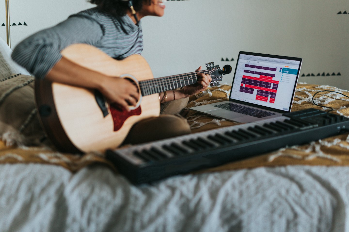 Person with headphones composes music on laptop with guitar and keyboard.